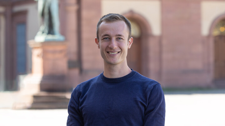 Lukas Kübek. He is wearing a blue sweater and is standing on the Ehrenhof in front of the castle.