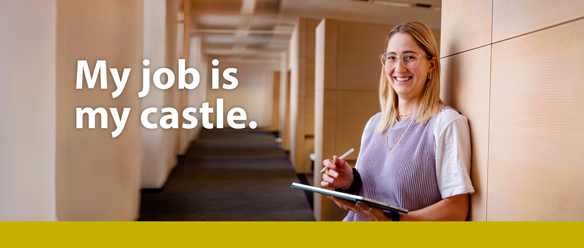 Foto mit Text: Eine junge Frau mit blonden Haaren und Brille lehnt an der Wand eines Uni-Flurs. In der Hand hält sie ein Tablet und einen Stift. Text: My job is my castle.