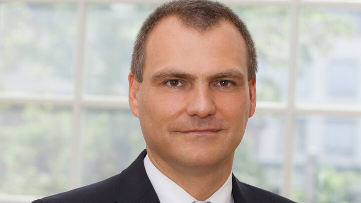 Portrait von Dirk Simons. Er trägt einen Anzug und steht vor einer Fensterfront.