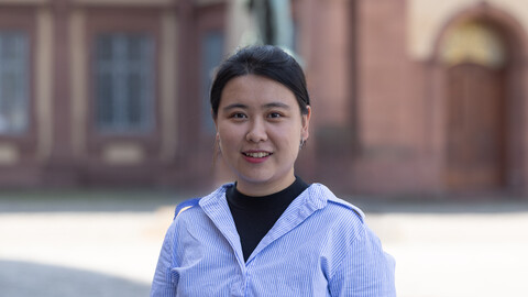 Yu Zhang. She is wearing a blue shirt over a black T-shirt and is standing on the Ehrenhof in front of the castle.