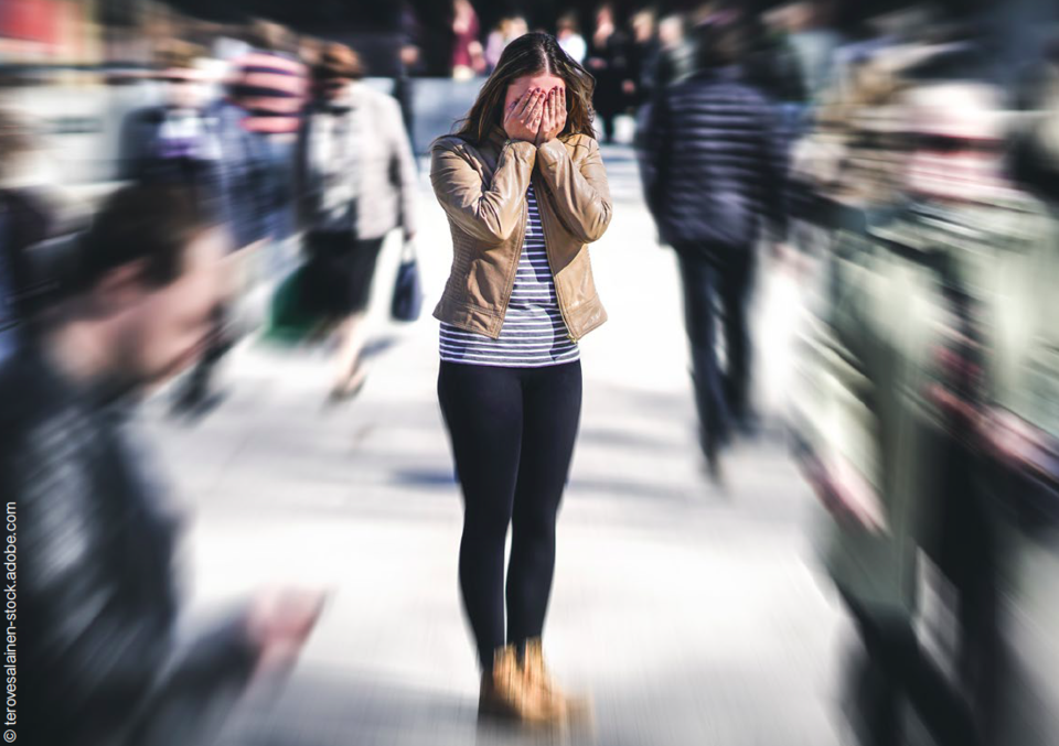 Vor einem Hintergrund mit Bewegungsunschärfe vieler undeutlicher Menschen in einem Fußgängerzonensetting eine junge Frau mit offenen dunklen Haaren, die sich die Hände vor ihr Gesicht hält. Sie träge eine beige Jacke über weißem Shirt mit dunklen Querstreifen sowie schwarze Hose und beige Schuhe.