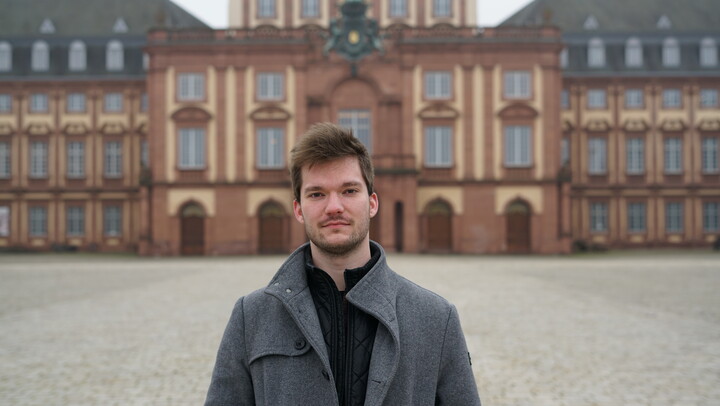 Picture of Julius Protte. He is wearing a grey jacket and standing in front of Mannheim Palace.