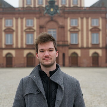 Picture of Julius Protte. He is wearing a gray jacket and standing in front of Schloss Mannheim.