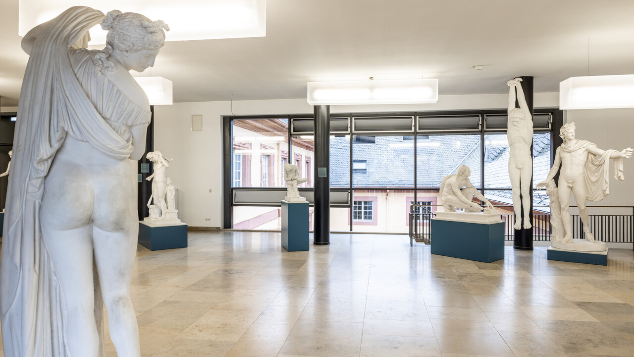 In einem Gang sind verschiedene Skulpturen von Menschen ausgestellt. Links im Vordergrund steht eine Skulptur einer Frau, die mit dem Rücken zur Kamera gedreht ist