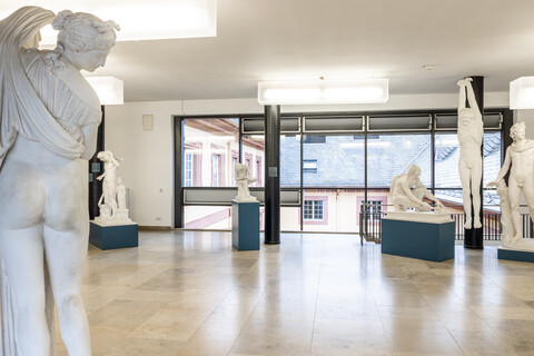 In einem Gang sind verschiedene Skulpturen von Menschen ausgestellt. Links im Vordergrund steht eine Skulptur einer Frau, die mit dem Rücken zur Kamera gedreht ist