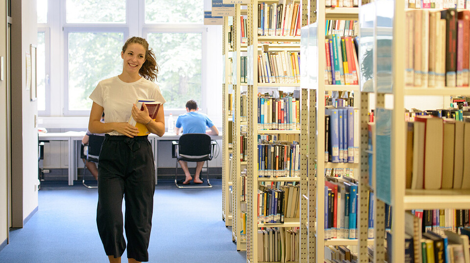 Eine Studentin läuft mit Büchern in der Hand durch eine Bibliothek. 