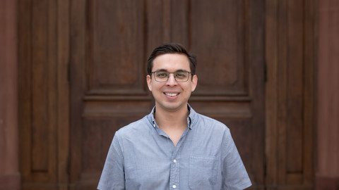 Andreas Lopez. He is wearing a grey shirt and is standing in front a brown door.