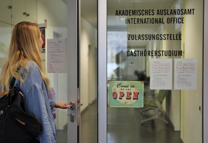 Eine junge Frau öffnet die Glastür des Akademischen Auslandsamtes der Universität Mannheim. Sie hat blonde Haare, trägt eine Jeansjacke und hält mit einer Hand die Türklinke fest.