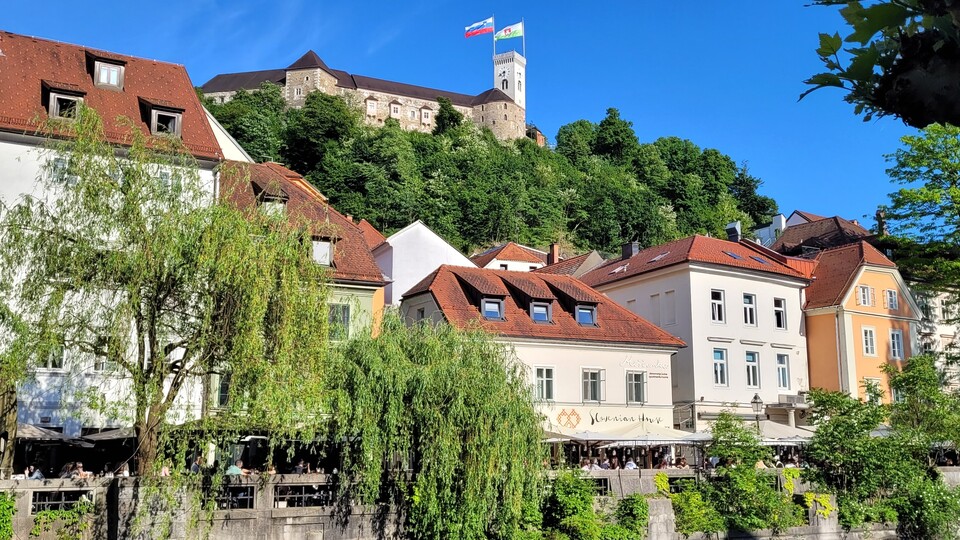 Blick auf einen Hügel mit bunten Gebäuden mit roten Dächern und einer üppigen Kulisse. Auf dem Hügel steht unter einem klaren blauen Himmel eine Burg, auf der zwei Fahnen wehen.