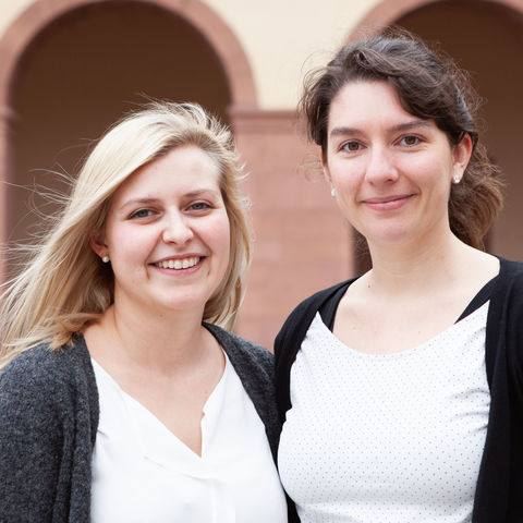 Zwei als Frauen gelesene Personen stehen lächelnd nebeneinander vor den Bogengängen des Ehrenhof West. Die Person links hat blondes Haar und trägt einen grauen Cardigan über einer weißen Bluse, die Person rechts hat braunes Haar, trägt einen schwarzen Cardigan über einem weißen Top mit Punkten.