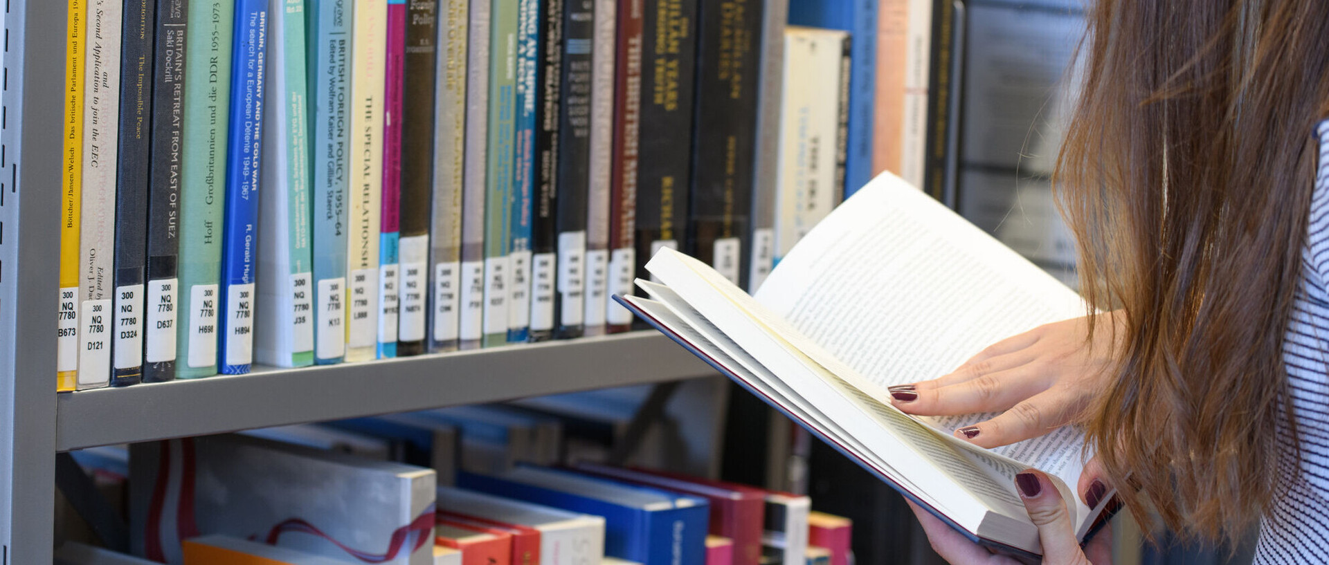 Eine Studentin steht in der Bibliothek vor einem Bücherregal und liest ein Buch.