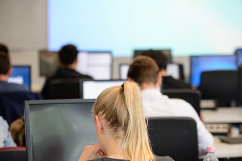 Rückenansicht von Studierenden, die in Reihen an Computer-Bildschirmen sitzen. 