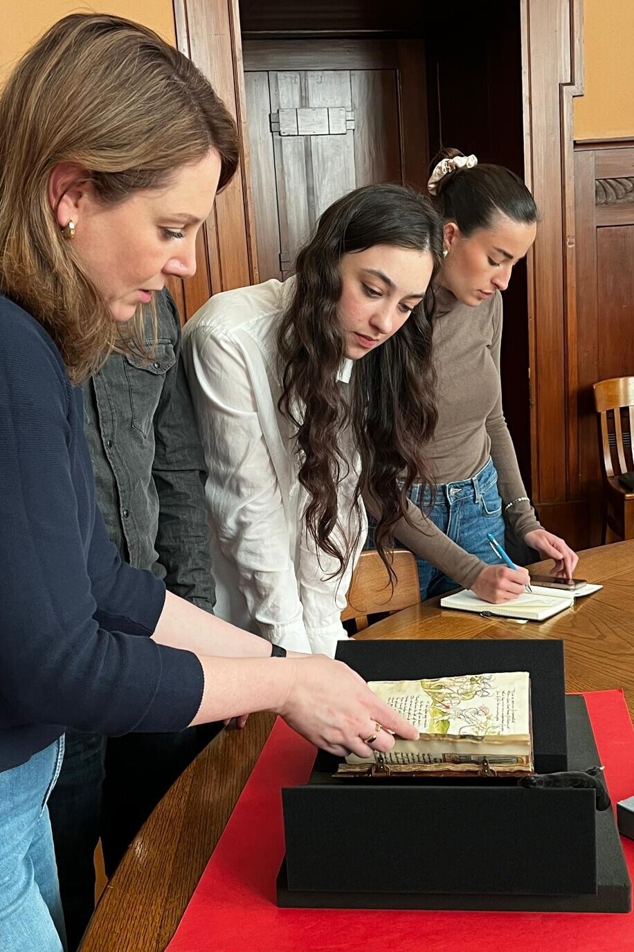 Eine Frau blättert in einer spätmittelalterlichen Handschrift, das auf einer Schaumstoffunterlage liegt, eine Frau sieht aufmerksam zu. Im Hintergrund macht sich eine andere Frau Notizen.