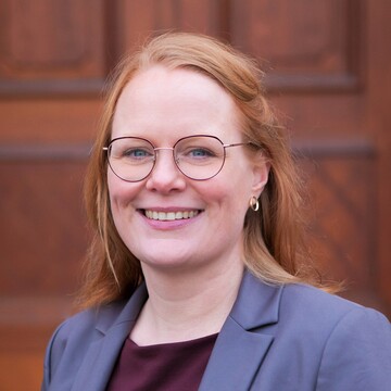 Solrun Graham-Parker hat rötliche, lange Haare. Sie trägt eine Brille und einen grauen Blazer.