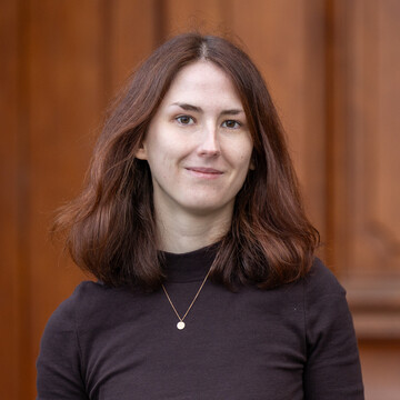 Portrait of Katharina Mengler. She is wearing a black sweater and standing in front of a door.