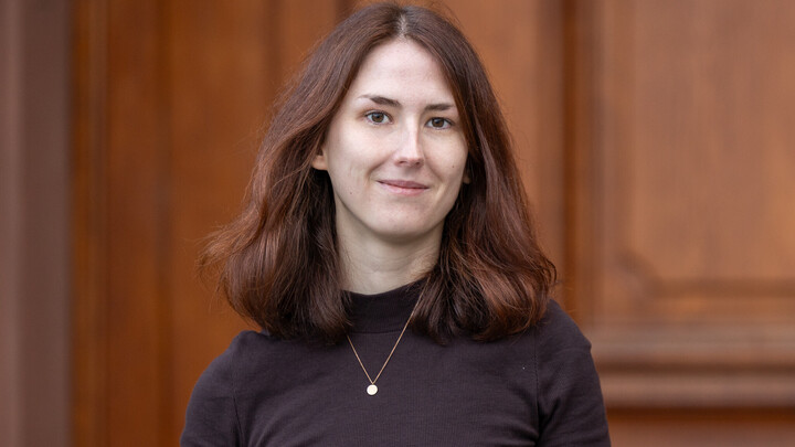 Portrait of Katharina Mengler. She is wearing a black sweater and standing in front of a door.