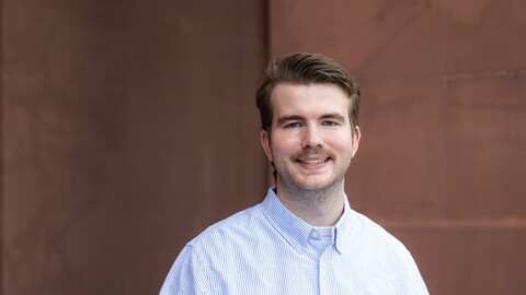 Lukas Warode. He is wearing a white shirt and is standing in front of a brown stone wall.