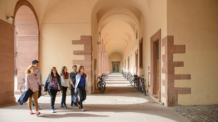 Vier Studentinnen und zwei Studenten spazieren über den Campus der Universität Mannheim. Die Studierenden sind frühlingshaft gekleidet. In einem Gang im Hintergrund stehen viele Fahrräder an den sandfarbenen Wänden des Schlosses.