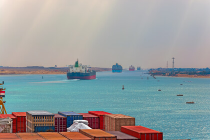 Im Vordergrund sind mehrere Container zu sehen. Im Hintergrund fahren durch einen Handelskanal mehrere Containerschiffe.