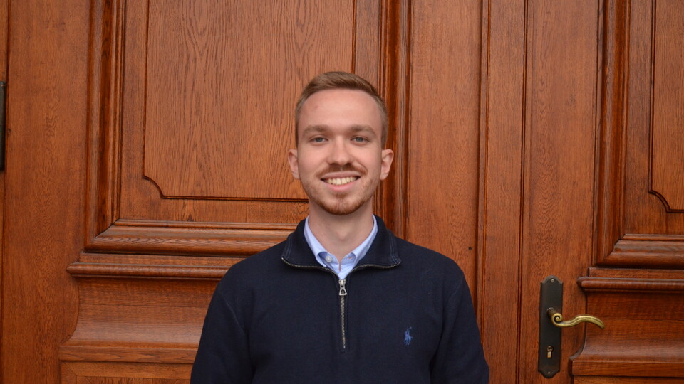 Maximilian Spanier vor der Eingangstür des Mittelbaus der Universität Mannheim. Er trägt einen dunkelblauen Pullover, darunter ein hellblaues Hemd und lächelt. 