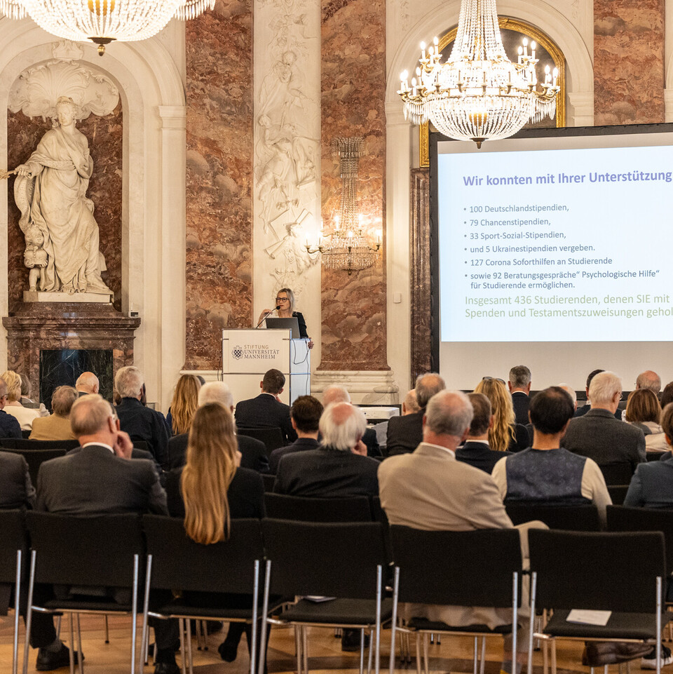 Mehrere Personen sitzen auf Stühlen und schauen auf eine Bühne, auf der Sabrina Scherbarth, ehrenamtliche Geschäftsführerin der Stiftung Universität Mannheim, eine Rede hält.