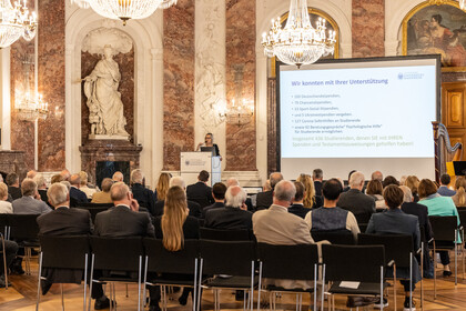 Mehrere Personen sitzen auf Stühlen und schauen auf eine Bühne, auf der Sabrina Scherbarth, ehrenamtliche Geschäftsführerin der Stiftung Universität Mannheim, eine Rede hält.