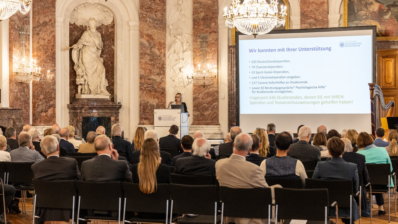 Mehrere Personen sitzen auf Stühlen und schauen auf eine Bühne, auf der Sabrina Scherbarth, ehrenamtliche Geschäftsführerin der Stiftung Universität Mannheim, eine Rede hält.