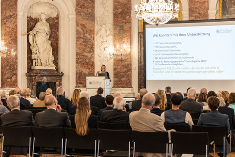 Mehrere Personen sitzen auf Stühlen und schauen auf eine Bühne, auf der Sabrina Scherbarth, ehrenamtliche Geschäftsführerin der Stiftung Universität Mannheim, eine Rede hält.