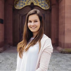 Fanni Szente hat braunes langes Haar und trägt eine weiße Bluse mit einer rosa Weste.Sie lächelt.