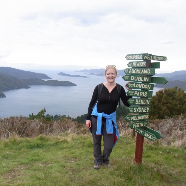 Eine Frau steht neben einem Schild mit Entfernungs- und Richtungsangaben verschiedener Städte der Welt. Im Hintergrund ein See mit Inseln und Bergen.