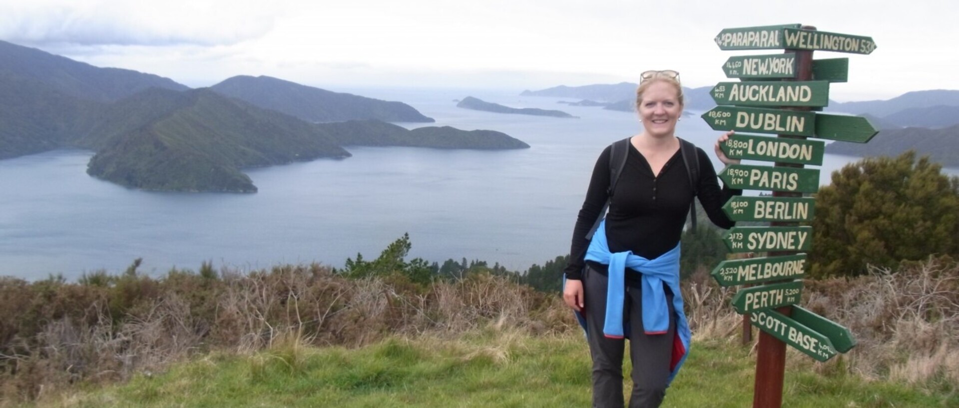 Eine Frau steht neben einem Schild mit Entfernungs- und Richtungsangaben verschiedener Städte der Welt. Im Hintergrund ein See mit Inseln und Bergen.