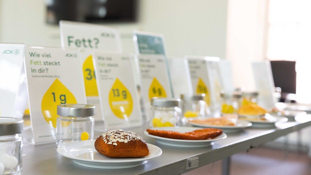 Teller auf einem Tisch mit verschiedenem Essen darauf. Dahinter Schilder mit den Fettangaben zu dem Produkt.