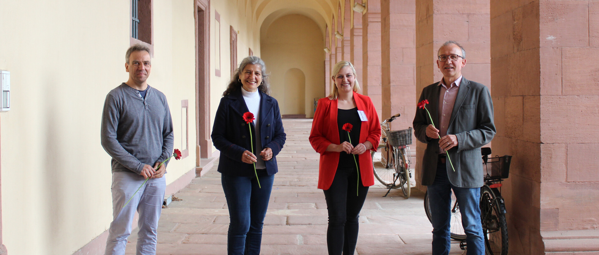 Vier Menschen stehen in einer Vorhalle des Mannheimer Schlosses. Alle lächeln und halten jeweils eine rote Blume.