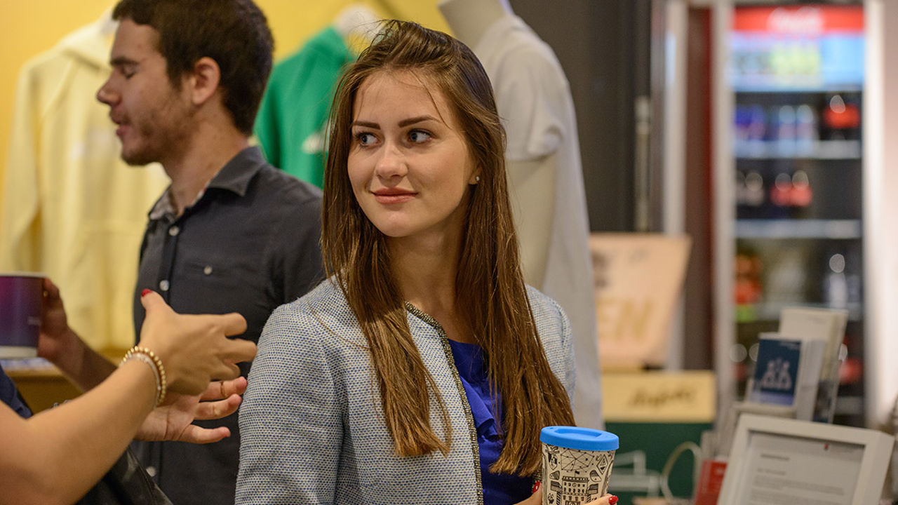 Eine Studentin hält einen wiederverwendbaren Kaffeebecher in ihrer Hand. Sie blickt nach rechts und trägt langes braunes Haar.