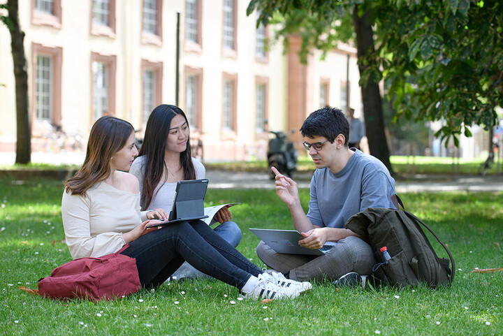 Three students are sitting on a meadow on campus. They have documents and laptops open in front of them.