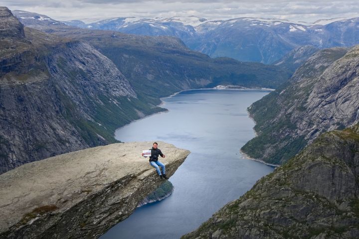 Ein junger Mann sitzt auf einem Felsen über einem Fjord in Norwegen. Er lächelt und hält ein Bild des Mannheimer Schlosses in seiner Hand.