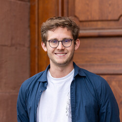 Vinzent trägt ein weißes T-Shirt und ein geöffnetes dunkelblaues Hemd. Er hat kurze hellbraune Haare, eine Brille und lächelt freundlich in die Kamera