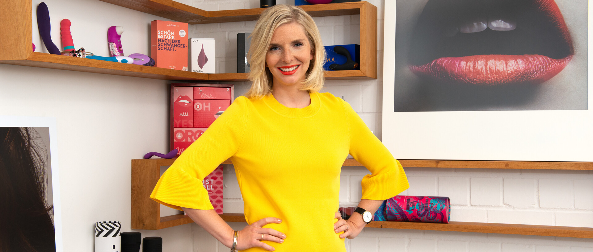Lea-Sophie Kramer trägt schulterlange blonde Haare, roten Lippenstift und ein knallgelbes Shirt. Sie hat die Arme in die Hüfte gestemmt und steht lächelnd vor einem Holzregal mit verschiedenen ihrer Produkte. Daneben hängt eine große Fotografie eines halb offenen roten Mundes.