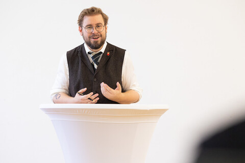 Eine Person trägt ein weißes Hemd sowie ein braunes Jackett und steht hinter einem Stehtisch und spricht. 