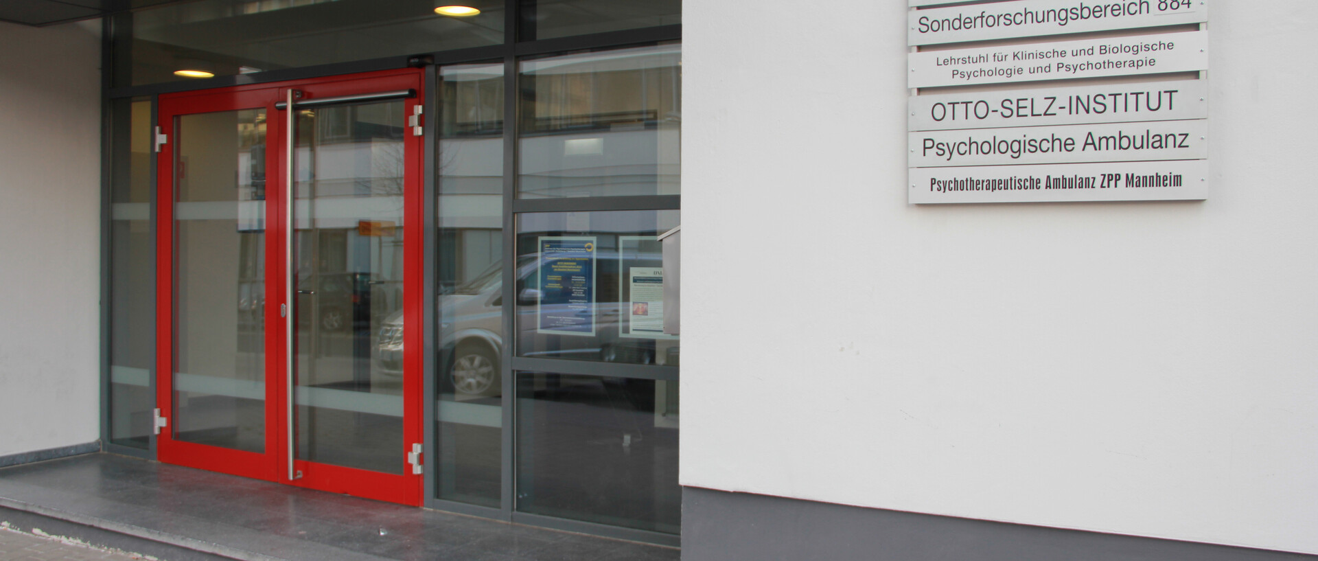 Zu sehen ist die rote Eingangstür des Otto-Selz-Instituts. Rechts davon befinden sich die Namen aller Lehrstühle und Institutionen, die im Gebäude vertreten sind (Psychologische Ambulanz...).