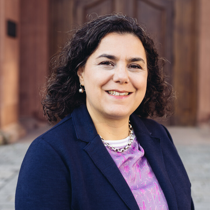 Portrait von Carmela Aprea. Sie trägt eine Bluse und einen dunkelblauen Blazer und steht vor dem Schloss der Uni Mannheim.