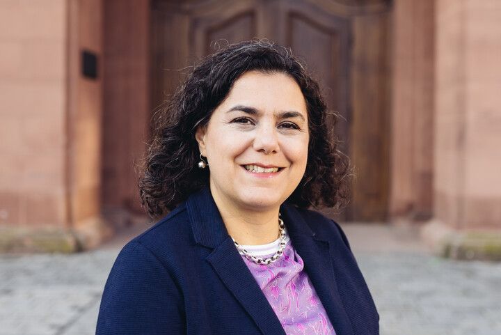 Portrait von Carmela Aprea. Sie trägt eine Bluse und einen dunkelblauen Blazer und steht vor dem Schloss der Uni Mannheim.
