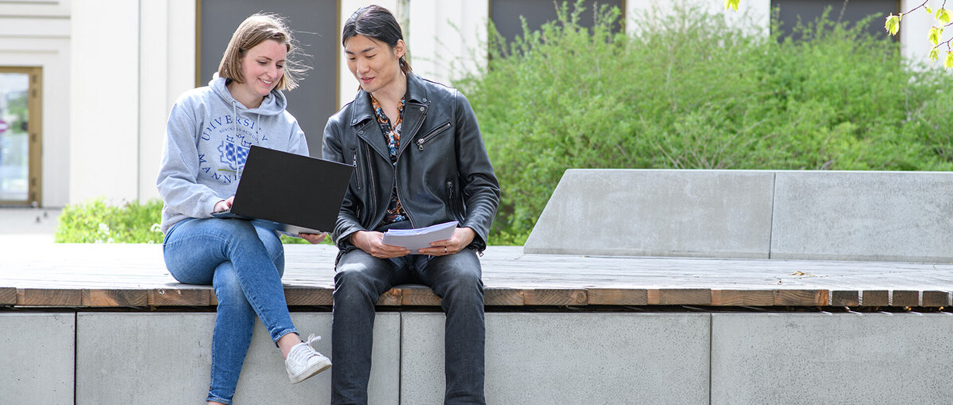 A woman and a man are sitting on a bank in the courtyard of the B6 building with a laptop and are discussing the content of the website on the screen. 
