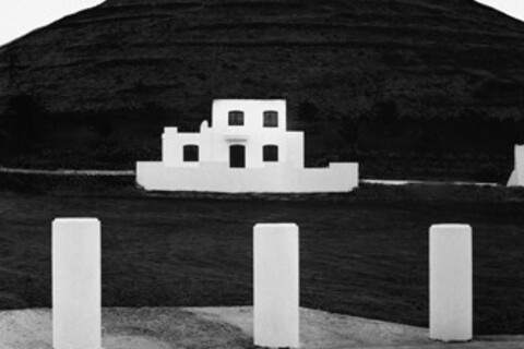 Schwarz-weiß Foto: vor einem großen Hügel ist ein weißes Haus. Im Vordergrund sind drei kleine Säulen.