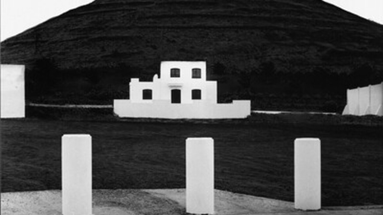 Schwarz-weiß Foto: vor einem großen Hügel ist ein weißes Haus. Im Vordergrund sind drei kleine Säulen.