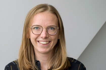 Fiona Draxler vor einer weißen Wand. Sie hat blonde Haare, trägt eine schwarze Bluse und eine goldene Brille.