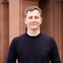 A portrait of Florian Braun in front of the red stone facade of the Schloss. He is grinning, has short blond hair, and is wearing a black sweater. He has his hands in the pockets of his blue jeans.
