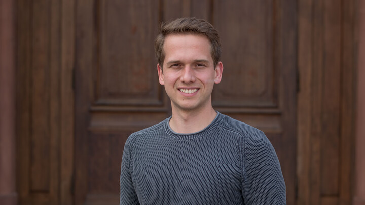 Johannes Gessner. He is wearing a grey sweater and is standing in front of a brown door.