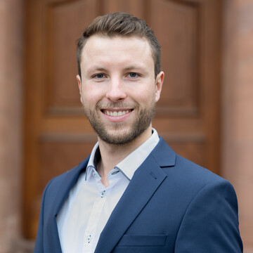 Philip Holler. Er trägt einen blauen Anzug und steht vor einer Holztür.