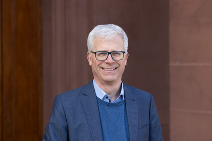 Portrait von Eckhard Janeba. Er trägt ein hellblaues Hemd, einen blauen Pullover sowie einen blauen Blazer und steht lächelnd vor einem Gebäude.